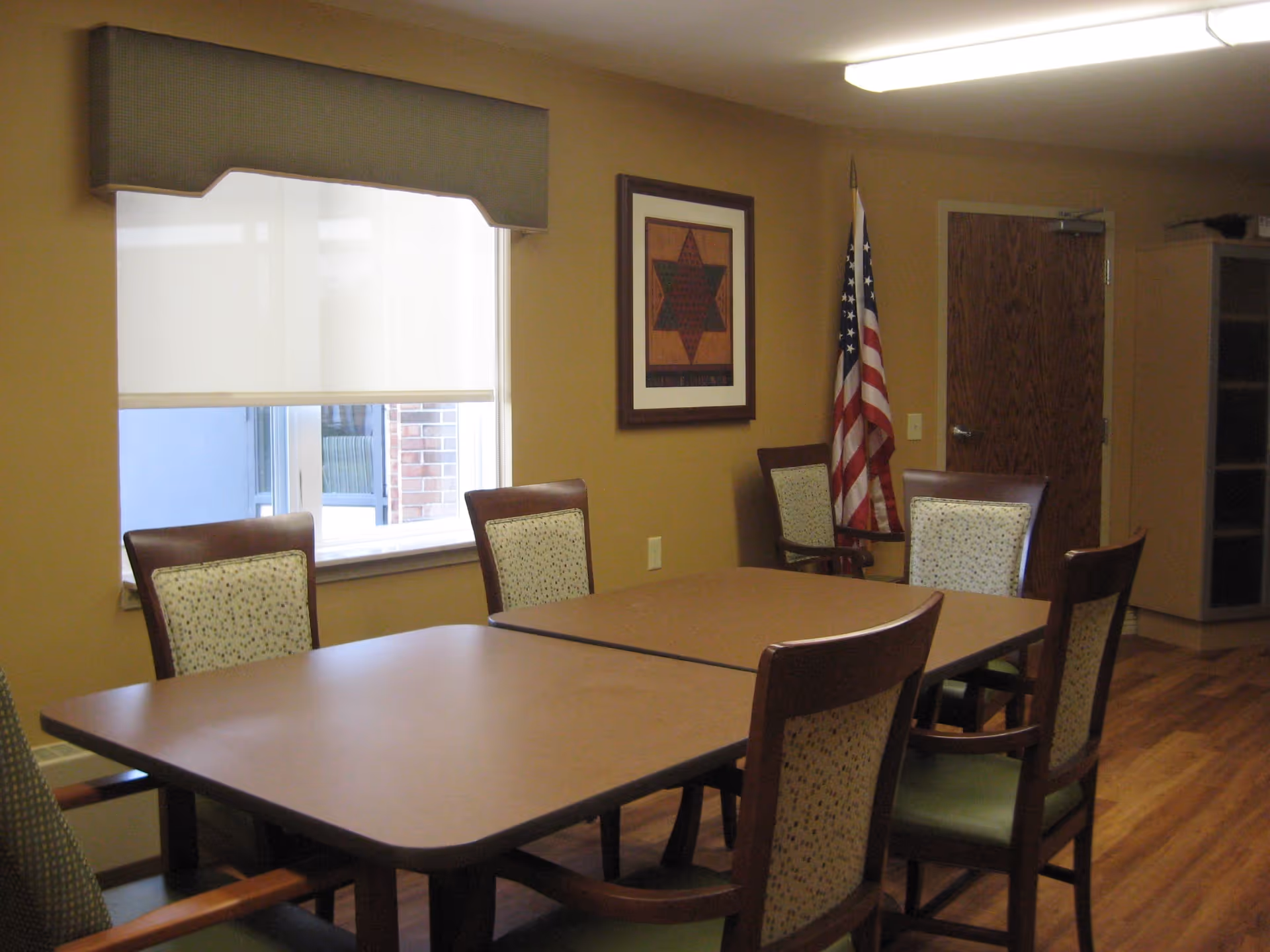 A room with two rectangular tables pushed together surrounded by six wooden chairs with patterned cushions. There is a window with a white roller shade and a valance, a framed artwork on the wall, an American flag in the corner, a wooden door, and a cabinet with glass doors. The floor is wood and the walls are painted beige.