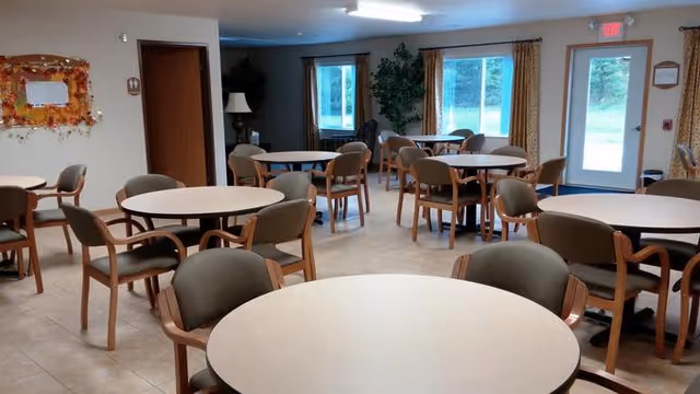 A bright dining room with multiple round tables and cushioned chairs arranged neatly. Large windows and a glass door allow natural light to enter, and there is a decorative fall-themed wreath on the wall. The room has a clean, welcoming atmosphere.
