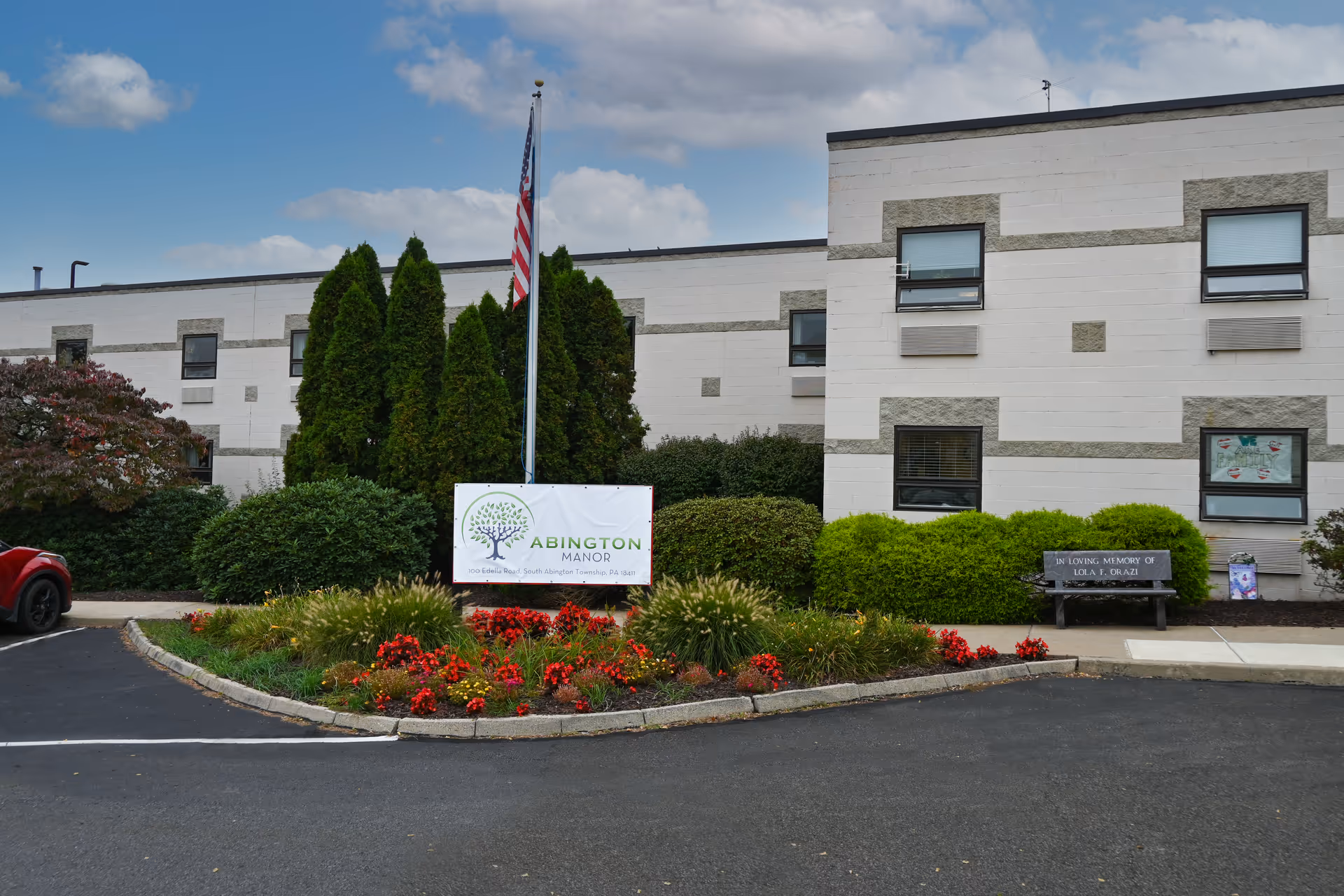 Exterior view of Abington Manor, a two-story building with white and gray brick walls, several windows, and landscaping including bushes and flowers. An American flag is displayed on a flagpole in front of the building, along with a sign that reads 'Abington Manor'. A memorial bench is visible on the right side of the image.