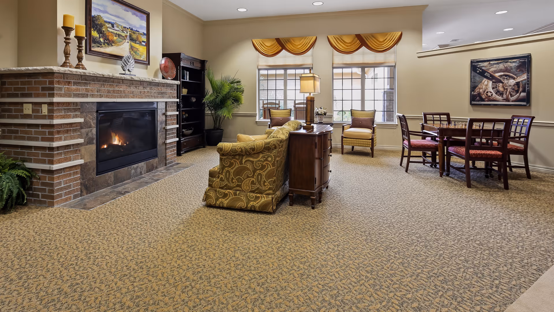 A cozy living room area with a lit brick fireplace on the left, a patterned sofa facing away from the fireplace, a wooden side table with a lamp, two armchairs near large windows with yellow valances, and a wooden table with four chairs on the right side. The room has beige walls and carpeted floor with a patterned design.