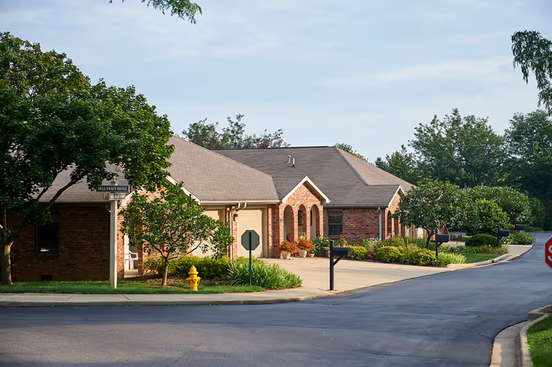 Exterior view of a single-story brick building with a sloped roof, surrounded by trees, shrubs, and a well-maintained driveway. There are mailboxes and a yellow fire hydrant near the curb, with a street sign that reads Bell Trace Drive.