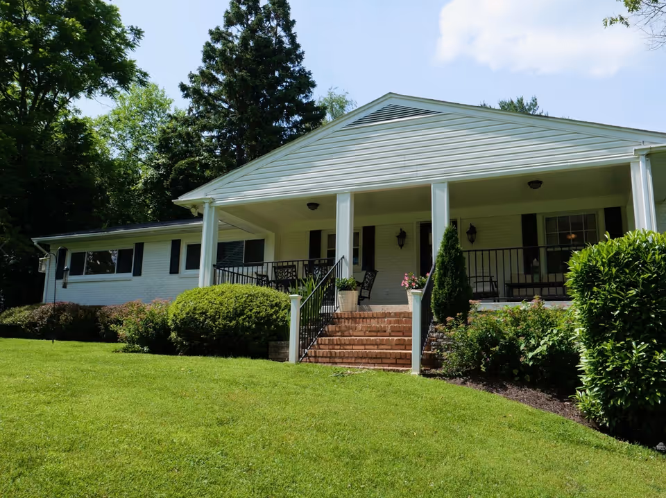 Front exterior view of a single-story white brick building with a covered porch, black railings, and outdoor seating. The building is surrounded by green bushes, trees, and a well-maintained lawn under a partly cloudy sky.