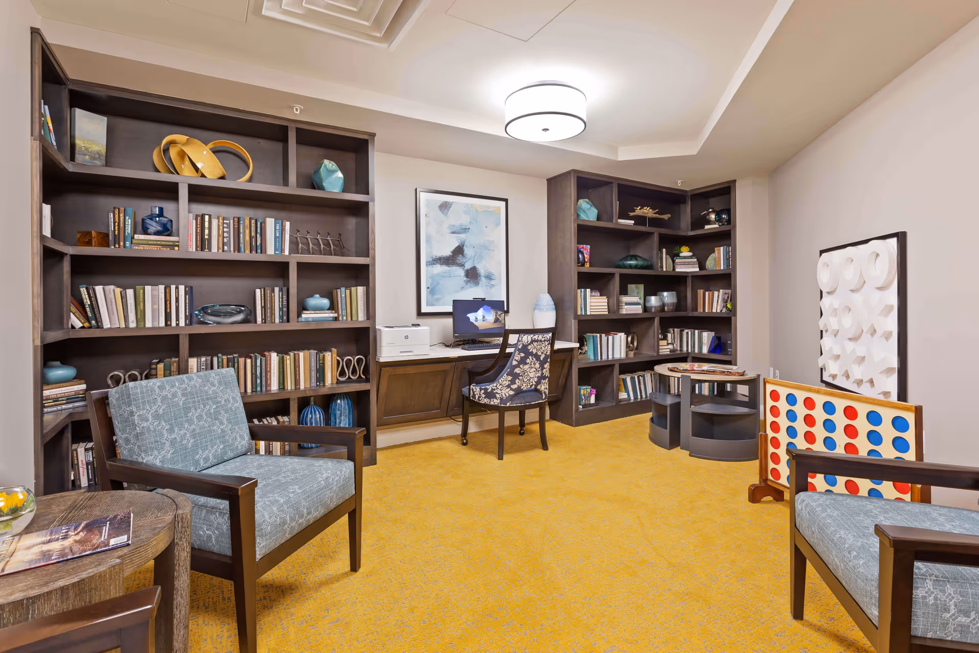 A cozy room with yellow carpet featuring two wooden armchairs with blue patterned cushions, dark wood bookshelves filled with books and decorative items, a desk with a computer and printer, and a wall-mounted tic-tac-toe game. The room is well-lit with a ceiling light and has a modern, inviting atmosphere.