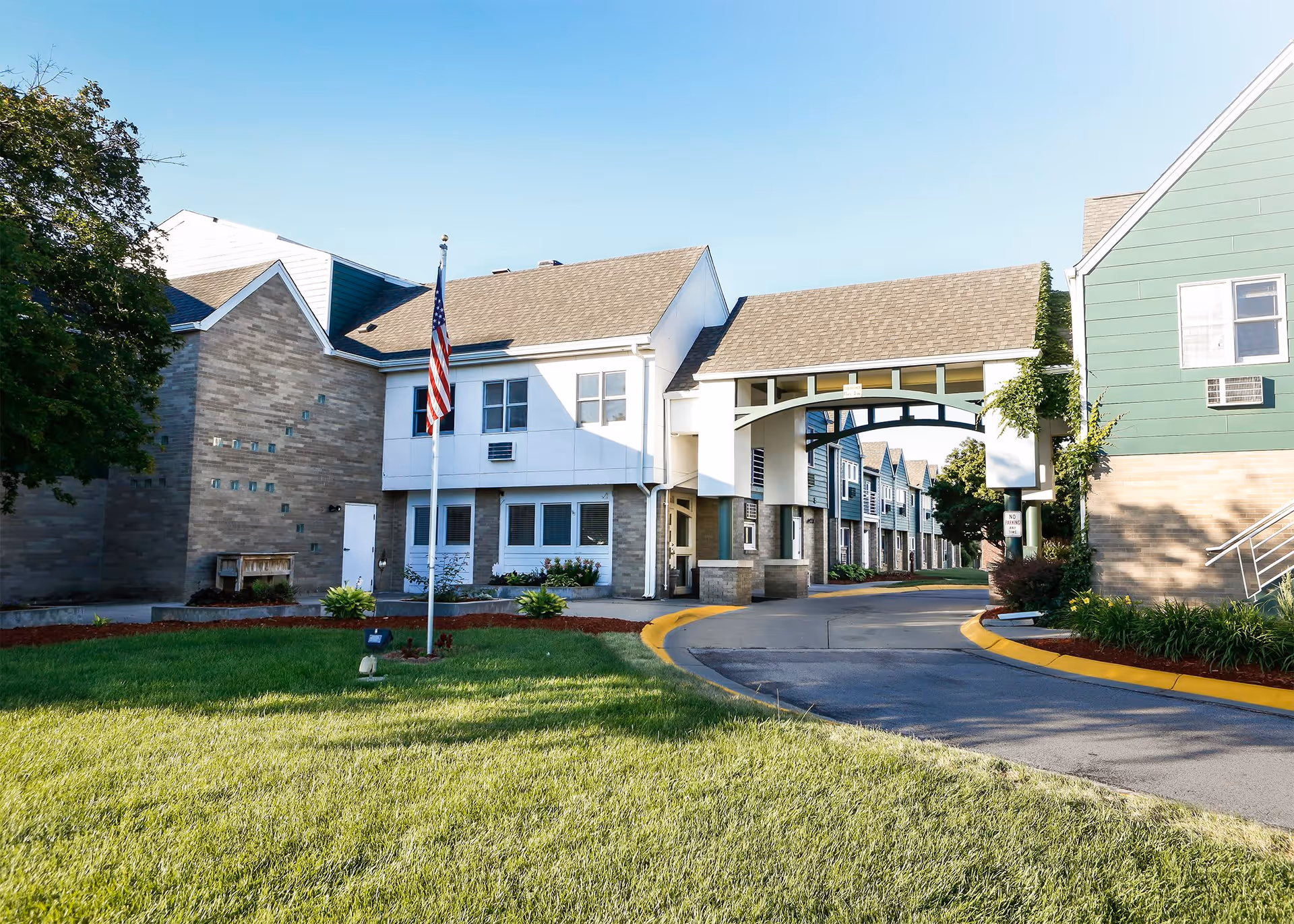 Exterior view of Hickory Villa Assisted Living facility showing a driveway with a covered archway connecting two buildings, green lawn with an American flag on a flagpole, and clear blue sky.