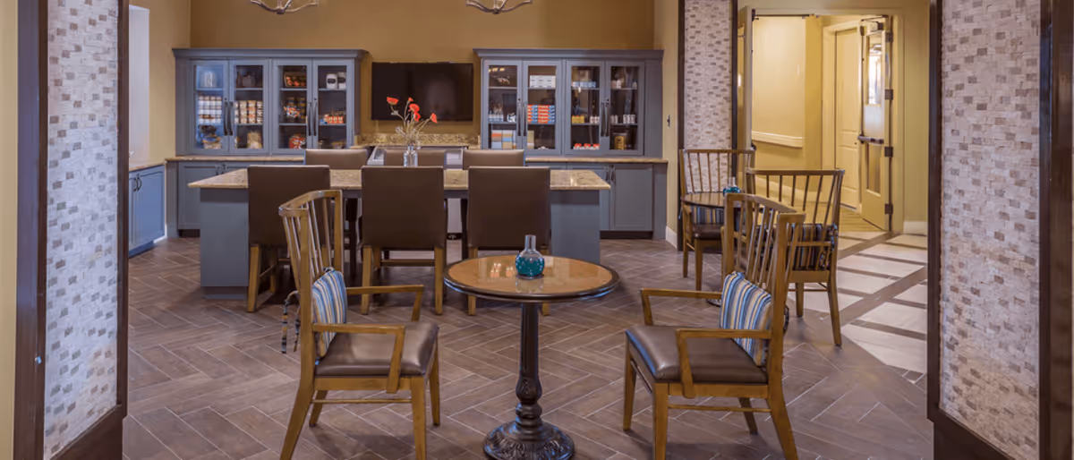 A cozy interior common area with a round wooden table and four wooden chairs with cushions in the foreground. In the background, there is a kitchen island with bar stools and blue cabinets with glass doors displaying various items. The room has warm lighting and a tiled floor with a herringbone pattern.