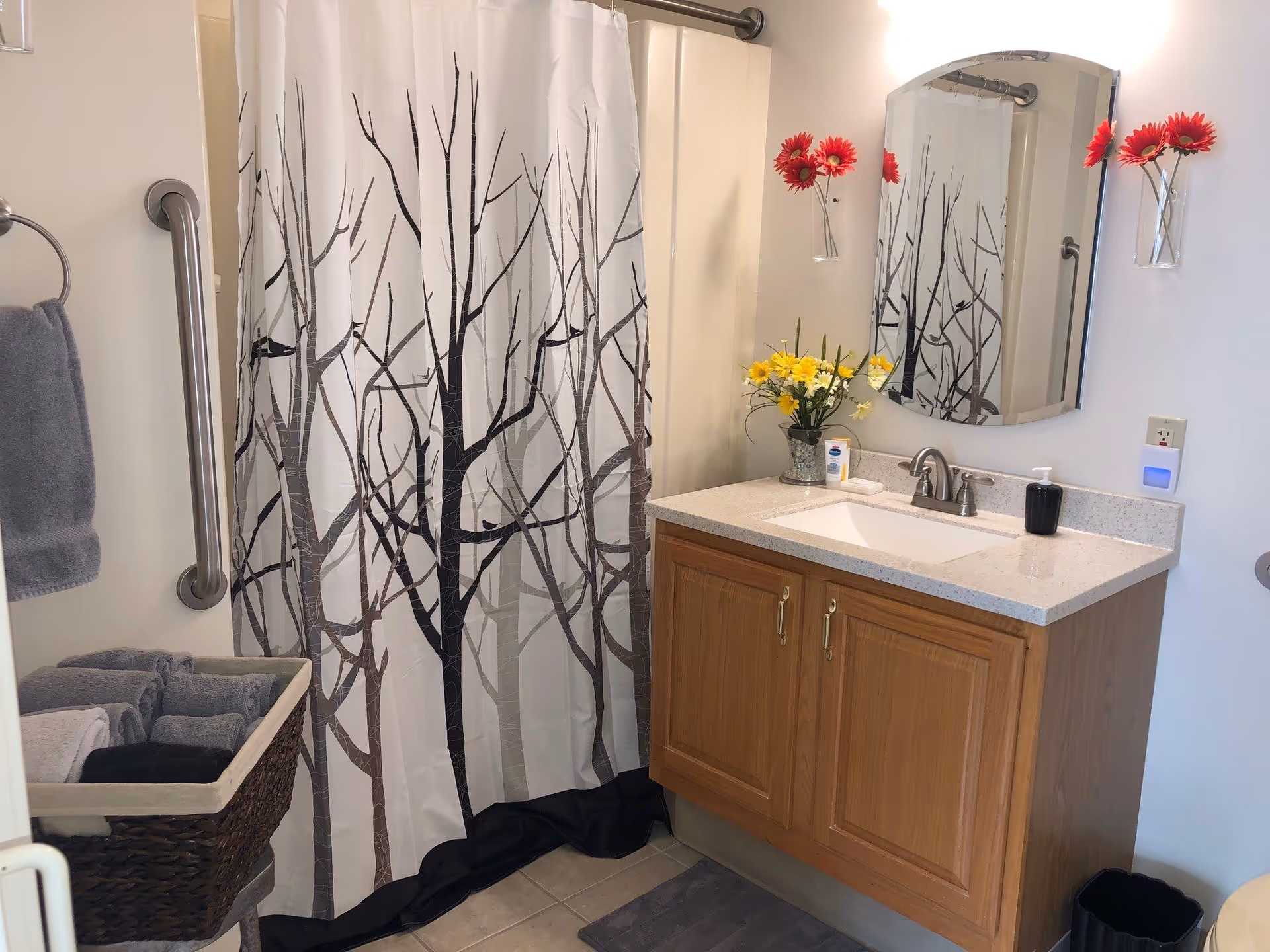 A bathroom with a wooden vanity cabinet topped with a white countertop and a sink. Above the sink is a mirror with two red flowers in wall-mounted glass vases on either side. To the left is a shower with a curtain featuring a tree branch design. A towel rack with a gray towel and a basket with folded towels are also visible.