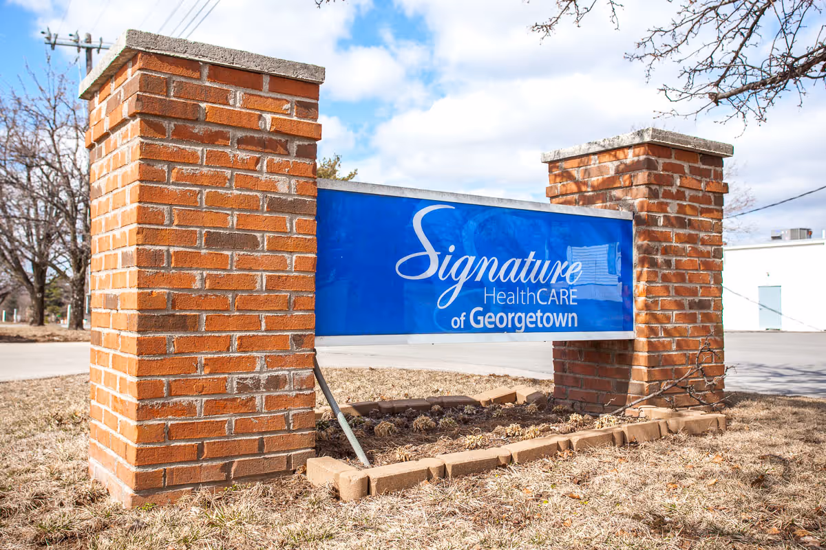 A blue sign reading "Signature HealthCARE of Georgetown" mounted between two brick pillars on a grassy verge.