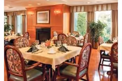 A warmly decorated dining room with several tables covered in beige tablecloths, each set with folded green napkins, plates, and cups. The room features wooden paneling, a fireplace, large windows with curtains, and upholstered chairs with floral patterns. There are plants near the windows adding a touch of greenery.