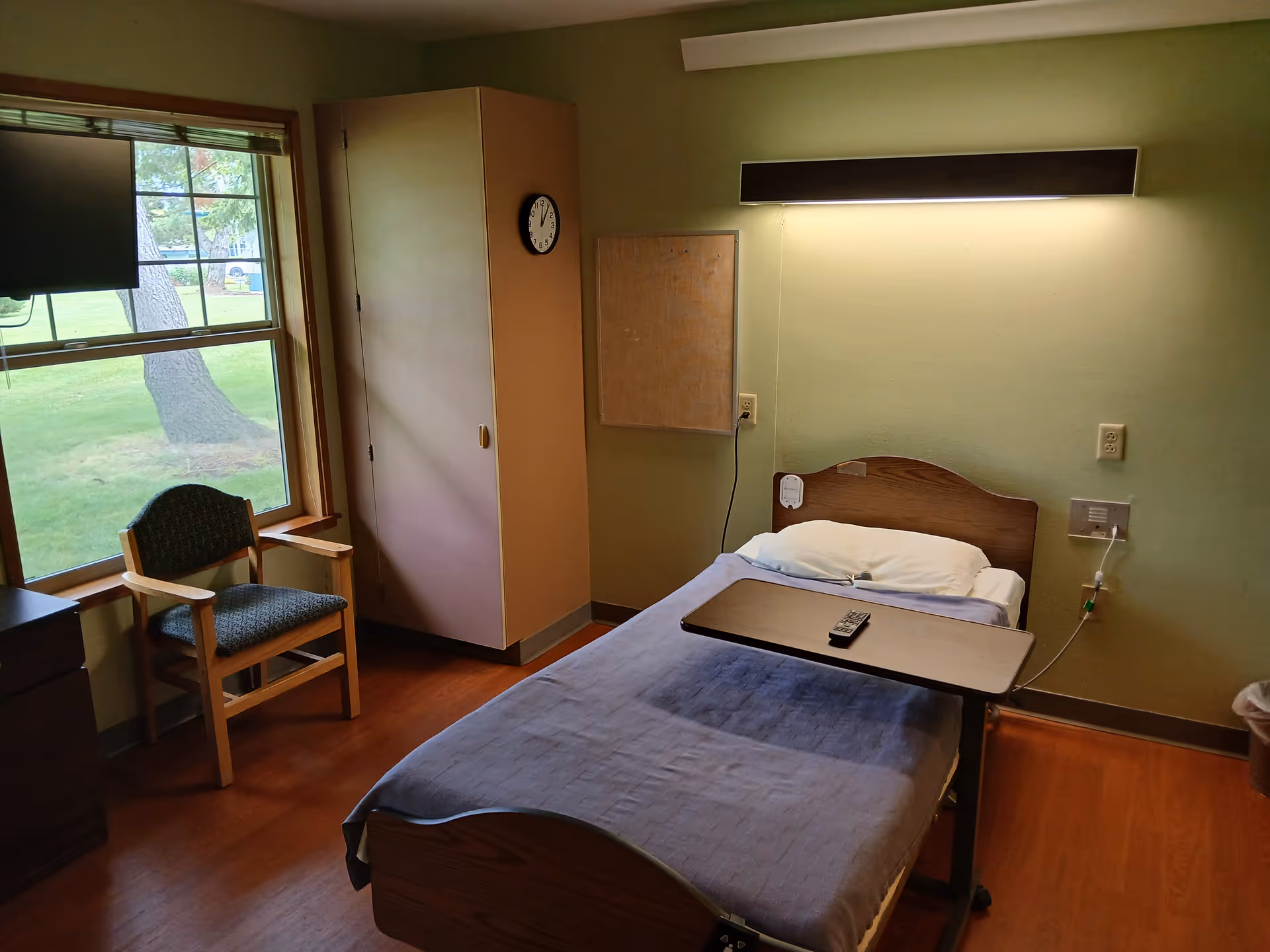 A simple nursing facility patient room with a single bed, overbed table and remote, a chair by a window, and a wardrobe against the wall.