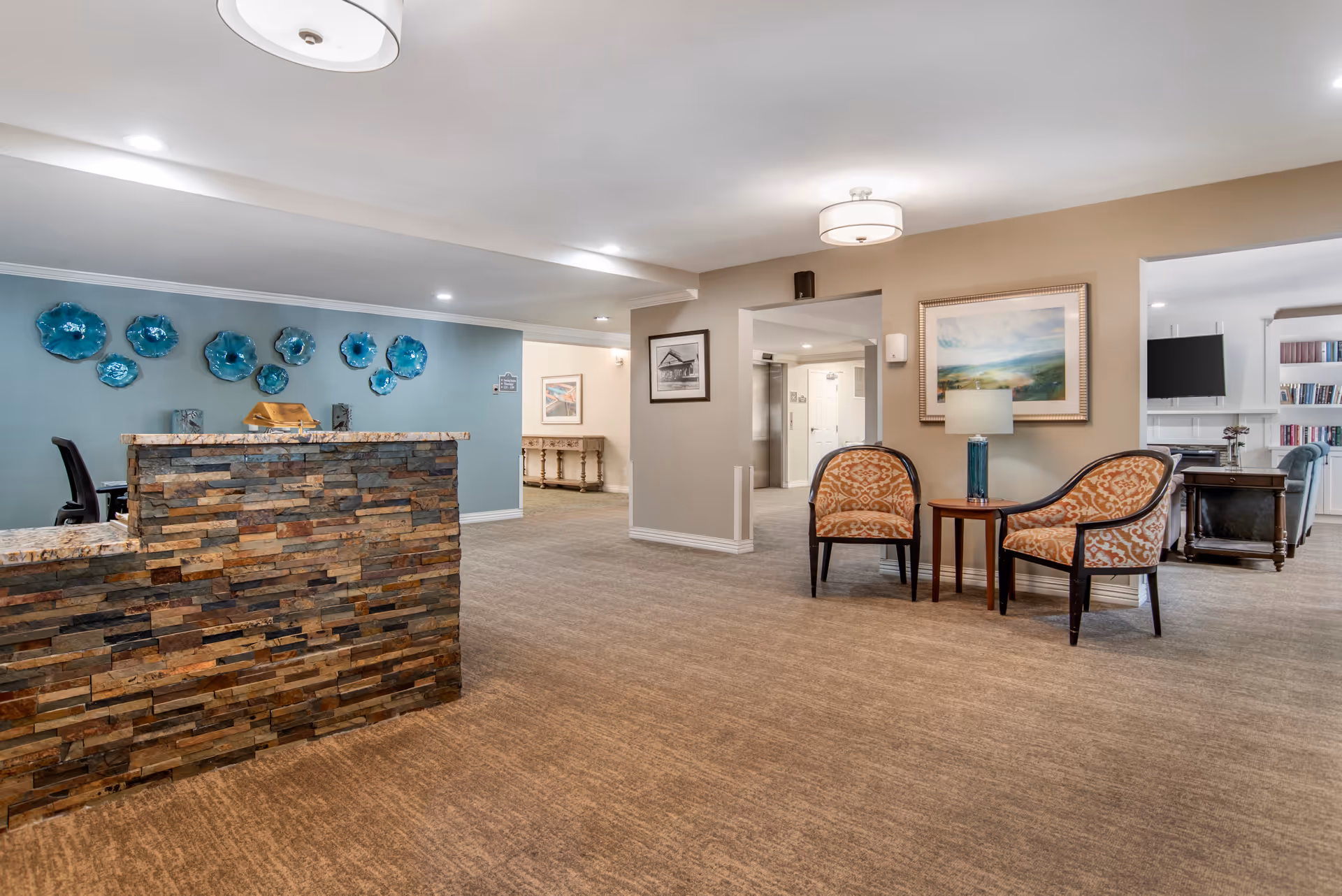 Interior view of a senior living facility lobby area with a stone reception desk on the left, blue decorative wall art behind the desk, two patterned chairs with a small table and lamp on the right, and a hallway leading to other rooms in the background.