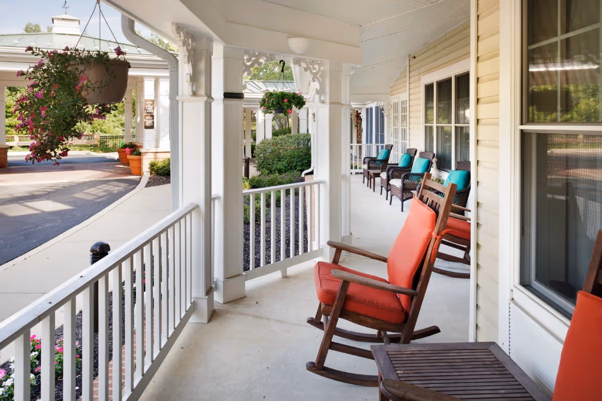 A covered porch area at Sunrise of Newtown Square with several wooden rocking chairs featuring red and teal cushions. Hanging flower pots and greenery decorate the space, with a paved driveway and garden visible beyond the porch.