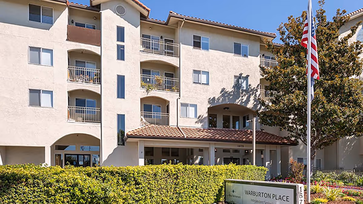Exterior view of Holiday Warburton Place, a multi-story senior living facility with balconies, a tiled roof entrance, an American flag on a flagpole, and well-maintained landscaping including bushes and trees.