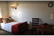 A simple, clean assisted living bedroom with a single bed made with white sheets, pillows, and a red blanket. There is a wooden chair next to a round table covered with a brown tablecloth, a lamp, and a telephone. A wall-mounted lamp and a clock are visible on the beige wall.