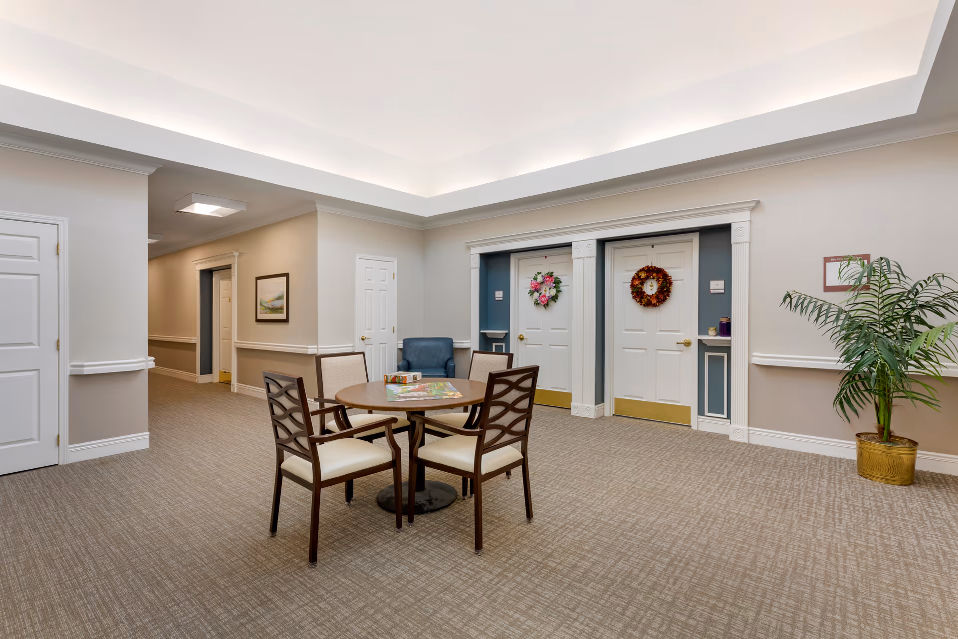 A well-lit common area in a senior living facility featuring a round table with four chairs around it. There is a board game on the table. The room has beige walls with white trim, two white doors decorated with wreaths, a blue armchair, a potted plant in a gold-colored pot, and a hallway leading to other rooms.