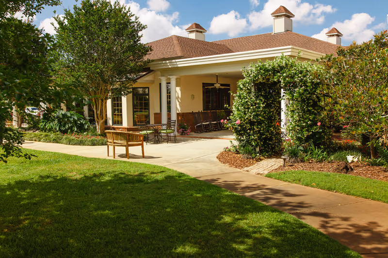 Outdoor patio area of a senior living facility with a covered porch featuring rocking chairs, a table with chairs, and a wooden bench. The area is surrounded by green grass, trees, and flowering bushes under a partly cloudy sky.