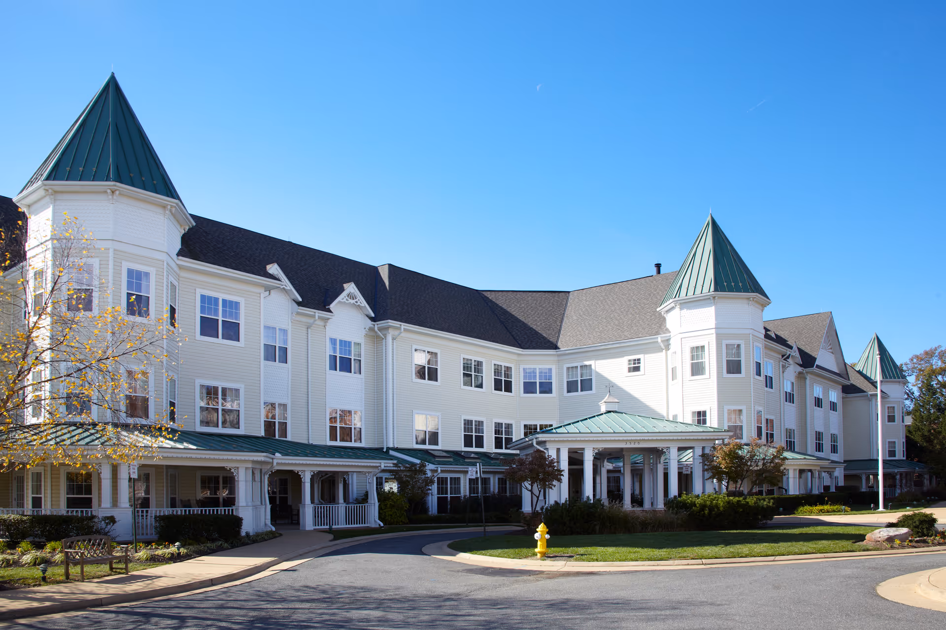 Exterior view of a large, multi-story senior living facility with white siding and green peaked roofs under a clear blue sky. The building has multiple windows and a covered entrance with columns. There is a circular driveway with a yellow fire hydrant and some landscaping including bushes and trees.