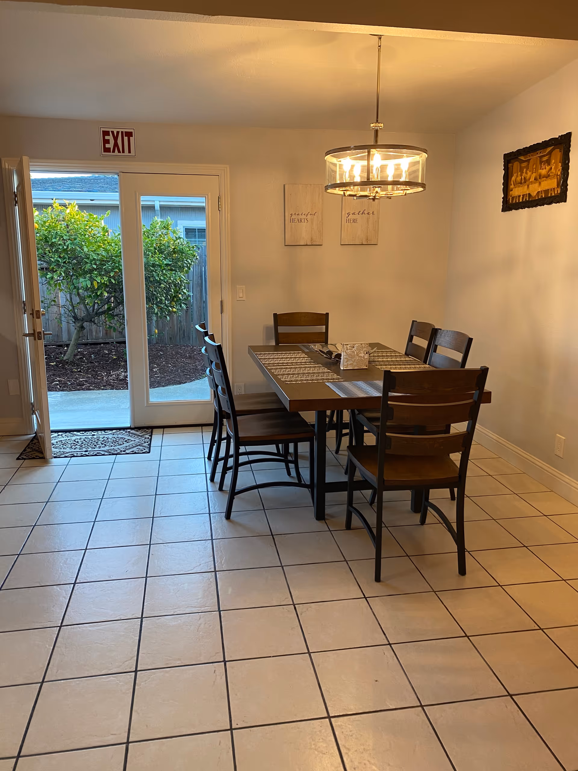 A dining area with a rectangular wooden table and six matching wooden chairs on a tiled floor. Above the table hangs a modern circular chandelier with multiple lights. On the wall behind the table are two decorative signs with text, and a framed picture. A glass door with an exit sign above it opens to an outdoor area with greenery and a wooden fence.