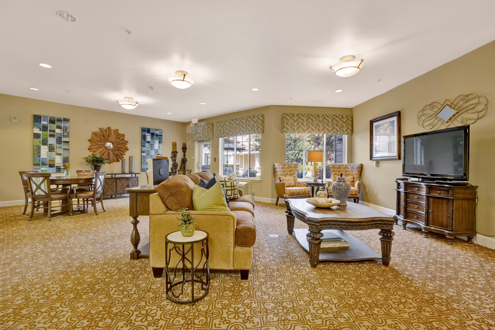 A spacious and well-lit living room area in a senior living facility featuring a beige sofa with colorful cushions, a wooden coffee table with decorative items, two armchairs near large windows with patterned valances, a flat-screen TV on a wooden cabinet, and a dining table with chairs in the background. The room has patterned carpet flooring and warm beige walls adorned with artwork and decorative mirrors.