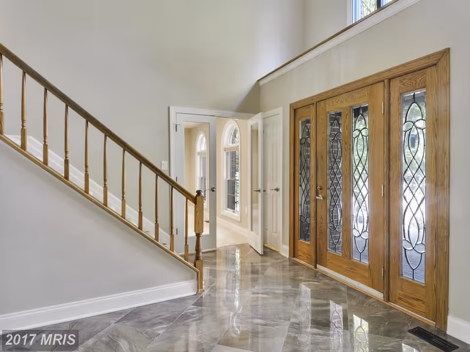 Interior view of a home's entryway featuring a wooden staircase with spindles on the left and a set of wooden double doors with decorative glass panels on the right. The floor is covered with shiny, reflective tiles, and there is a hallway with arched windows visible through an open door.