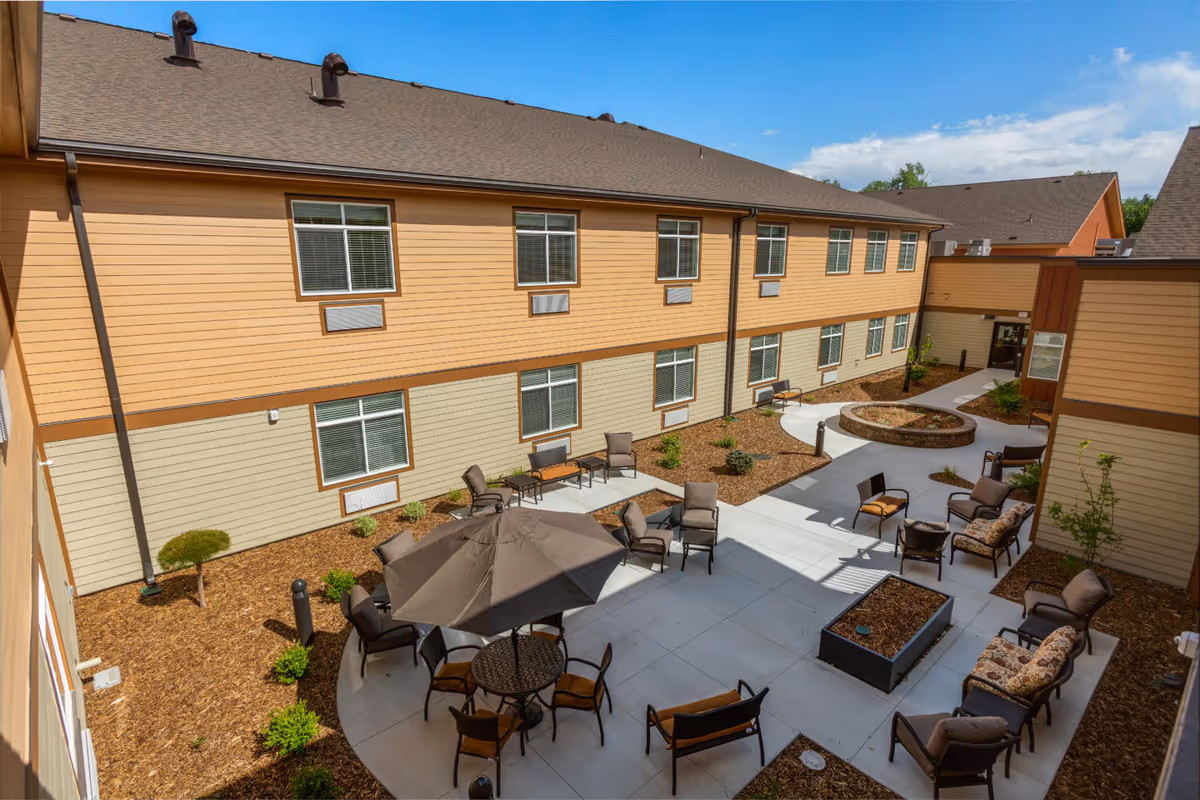 Outdoor courtyard area of a senior living facility with multiple seating arrangements including chairs, benches, and tables with umbrellas. The courtyard is surrounded by a two-story building with beige and light brown siding and several windows. There are small landscaped areas with mulch and plants, and a clear blue sky overhead.