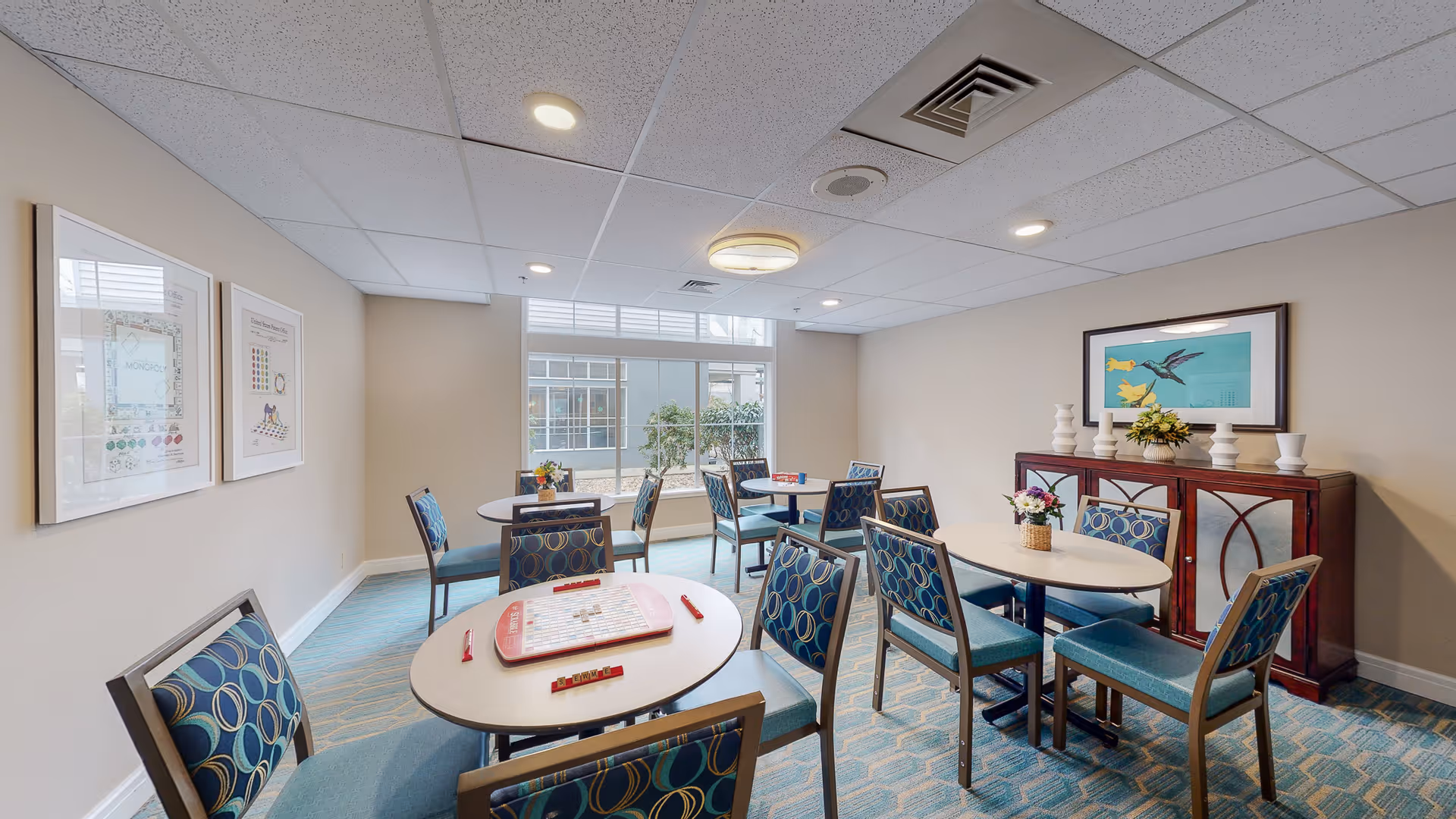 A well-lit activity room with multiple round tables and chairs arranged around them. One table has a Scrabble game set up. The room has a large window with a view of some greenery outside. The walls are decorated with framed artwork and a wooden cabinet with vases and a flower arrangement on top.