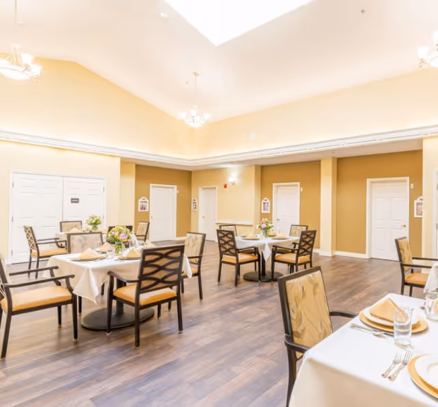 Bright, well-lit dining room with multiple set tables, chairs, and floral centerpieces under a high ceiling.