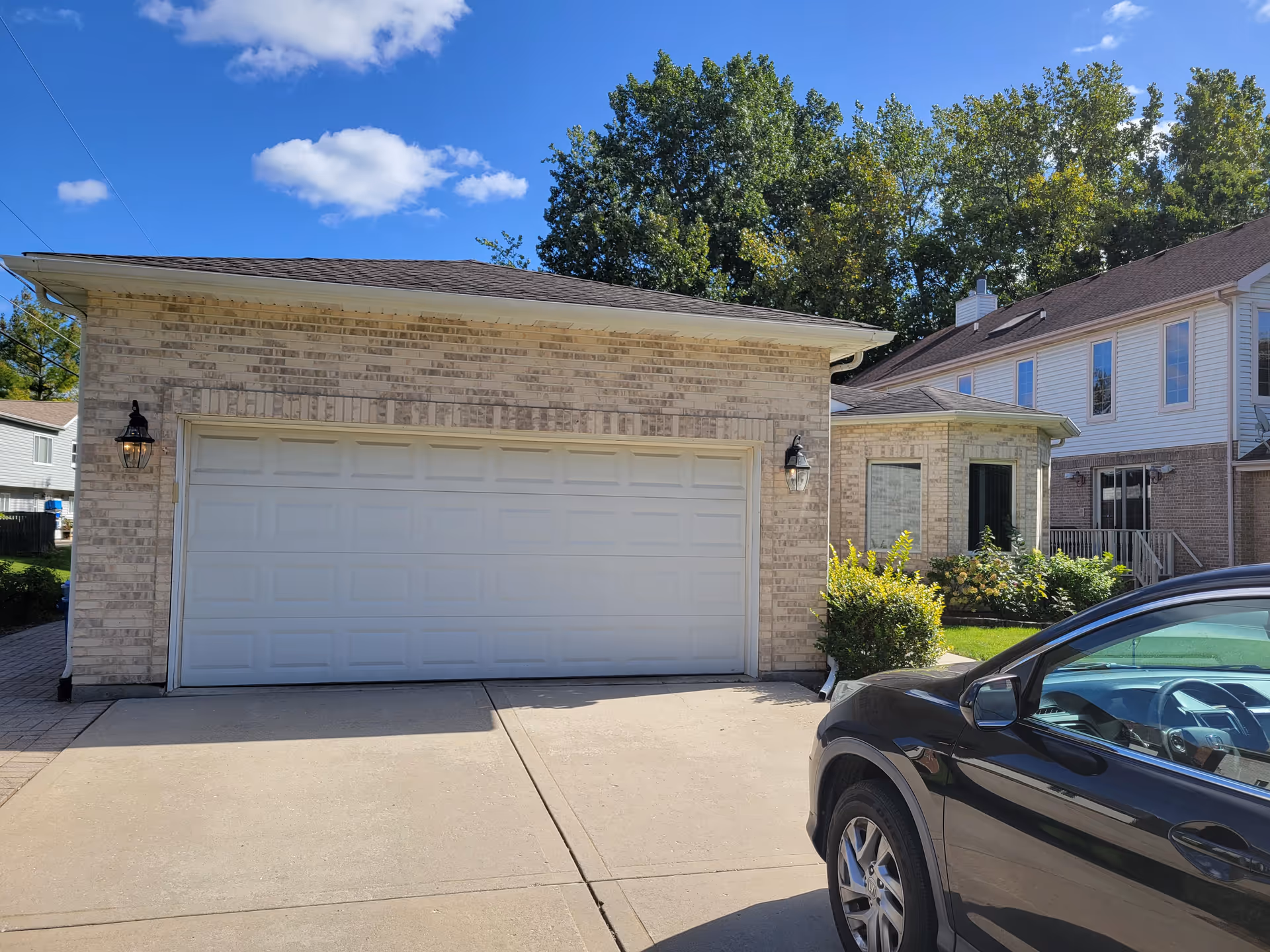 Exterior view of a residential garage with a closed white garage door, two wall-mounted lantern-style lights on either side, a black car parked on the driveway, and a house with windows and greenery in the background under a blue sky with some clouds.