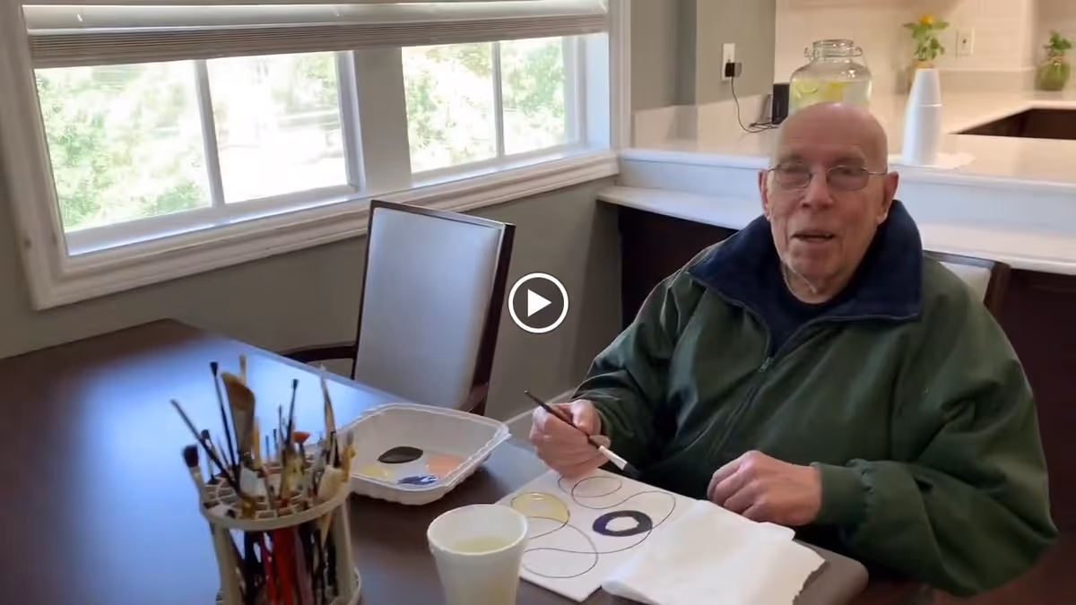 An elderly man wearing glasses and a green jacket is sitting at a table in a well-lit room with large windows. He is holding a paintbrush and painting a design with circles on a piece of paper. On the table, there is a container holding various paintbrushes, a palette with paint, and a cup of water. The room has a countertop with a glass jar and a small plant in the background.