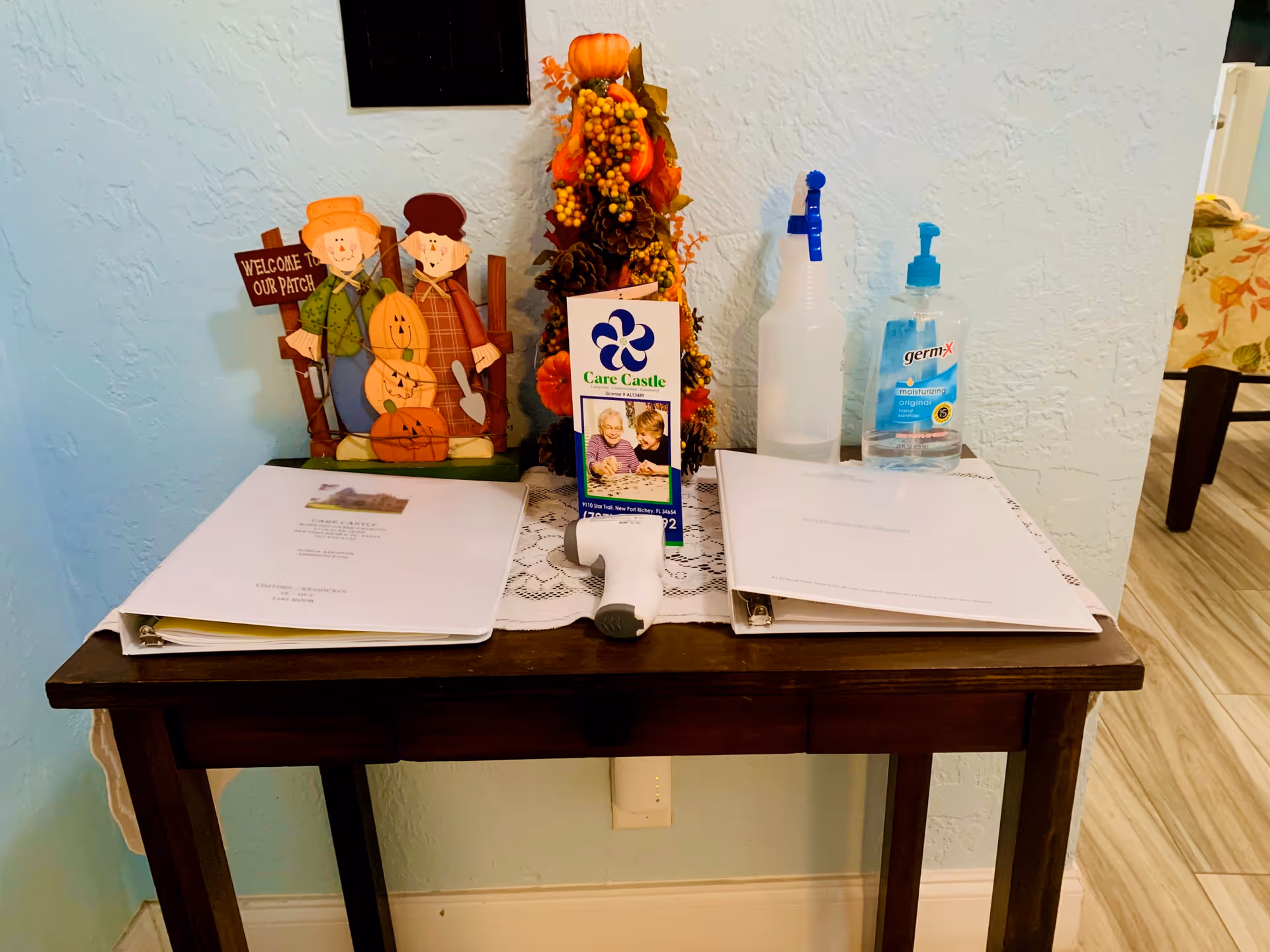 Small wooden table against a pale blue wall holding binders, a Care Castle brochure, hand sanitizer, a spray bottle, and fall decorations.
