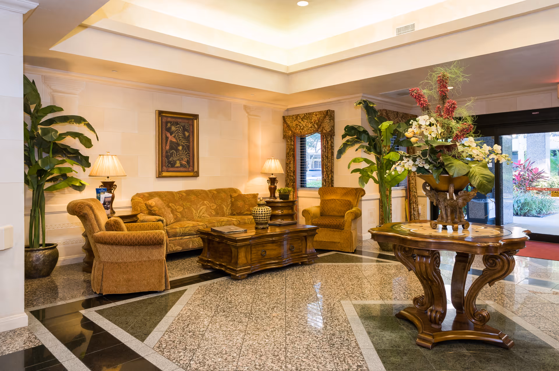 A well-lit, elegant lobby area with a patterned tile floor, a wooden coffee table surrounded by upholstered armchairs and a sofa, potted plants, table lamps, a framed painting on the wall, and a round wooden table with a large floral arrangement near the entrance door.