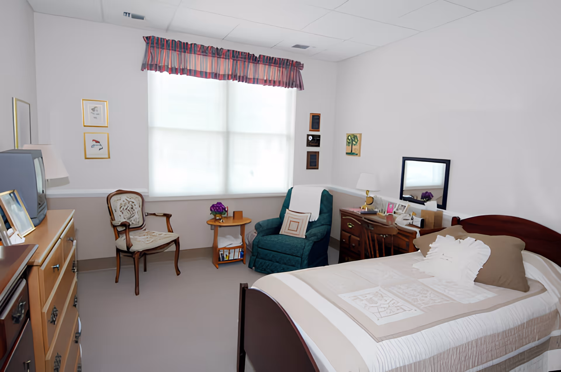 A cozy bedroom in a senior living facility featuring a single bed with beige and white bedding, a wooden dresser with a TV on top, a wooden chair with a cushioned seat, a green armchair with a pillow, a small round wooden side table with a purple flower pot, and a wooden desk with a mirror and various personal items. The room has a large window with sheer white curtains and a striped valance.