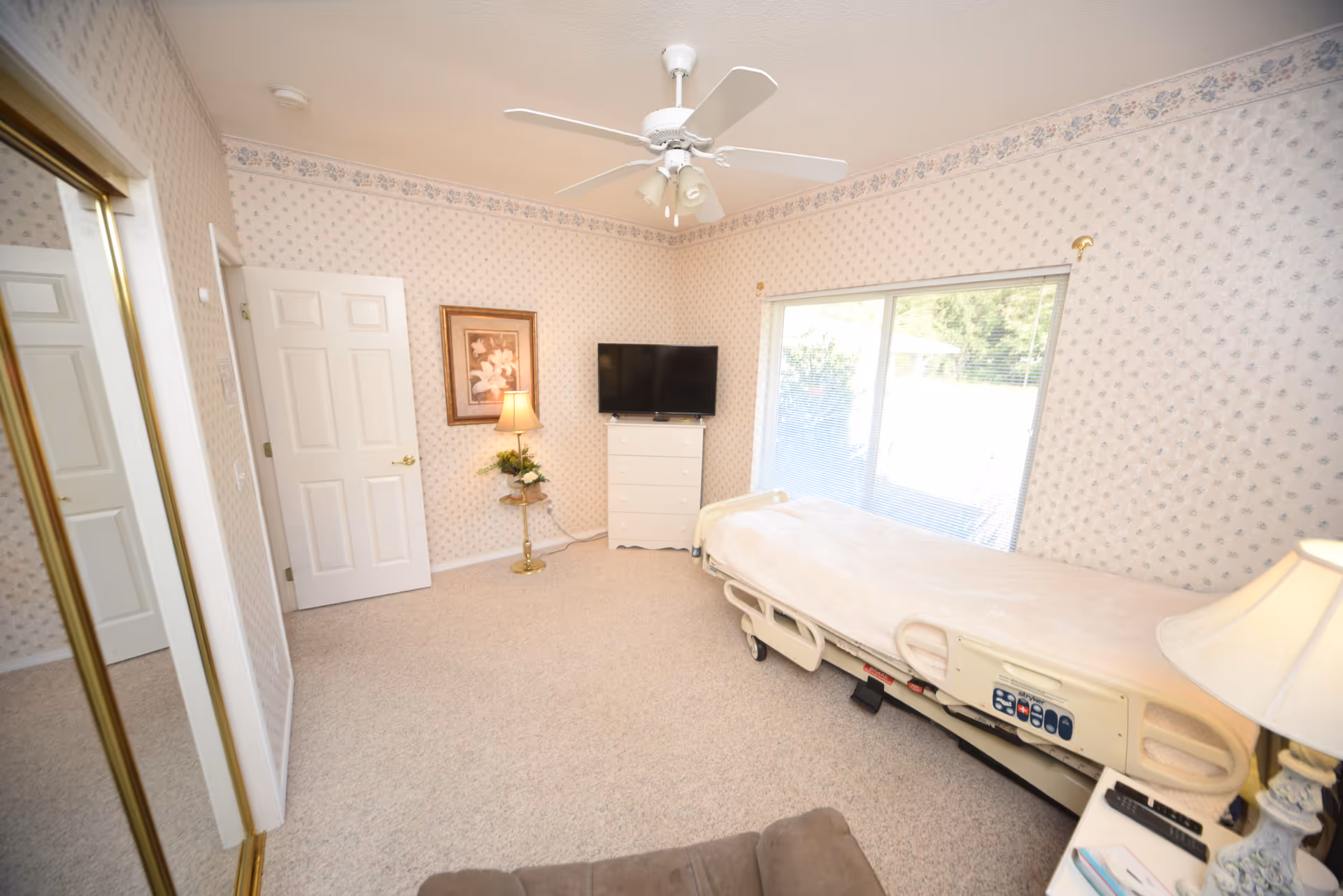 A bedroom in an adult family home featuring a hospital-style bed with beige bedding, a white dresser with a flat-screen TV on top, a floor lamp next to a framed floral picture on the wall, a ceiling fan with lights, a sliding glass door with blinds, and a mirrored closet door reflecting part of the room.