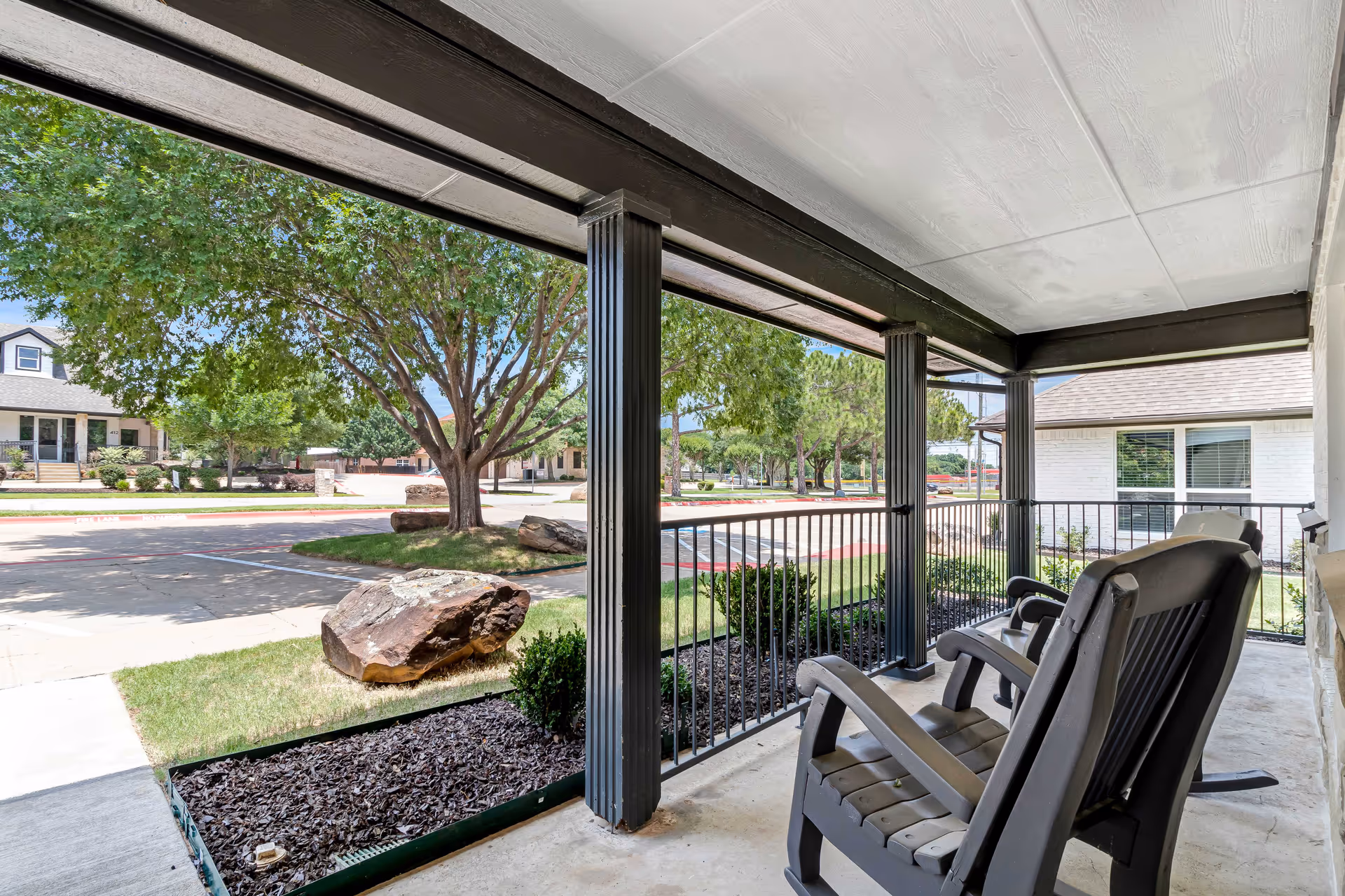 Covered porch area with two black rocking chairs facing outward. The porch overlooks a landscaped area with mulch, bushes, large rocks, and a tree. Across the street, there are residential-style buildings and more trees under a clear blue sky.