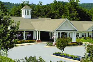 Front entrance of a single-story senior living building with a covered porte-cochère, circular driveway and landscaped flower beds.