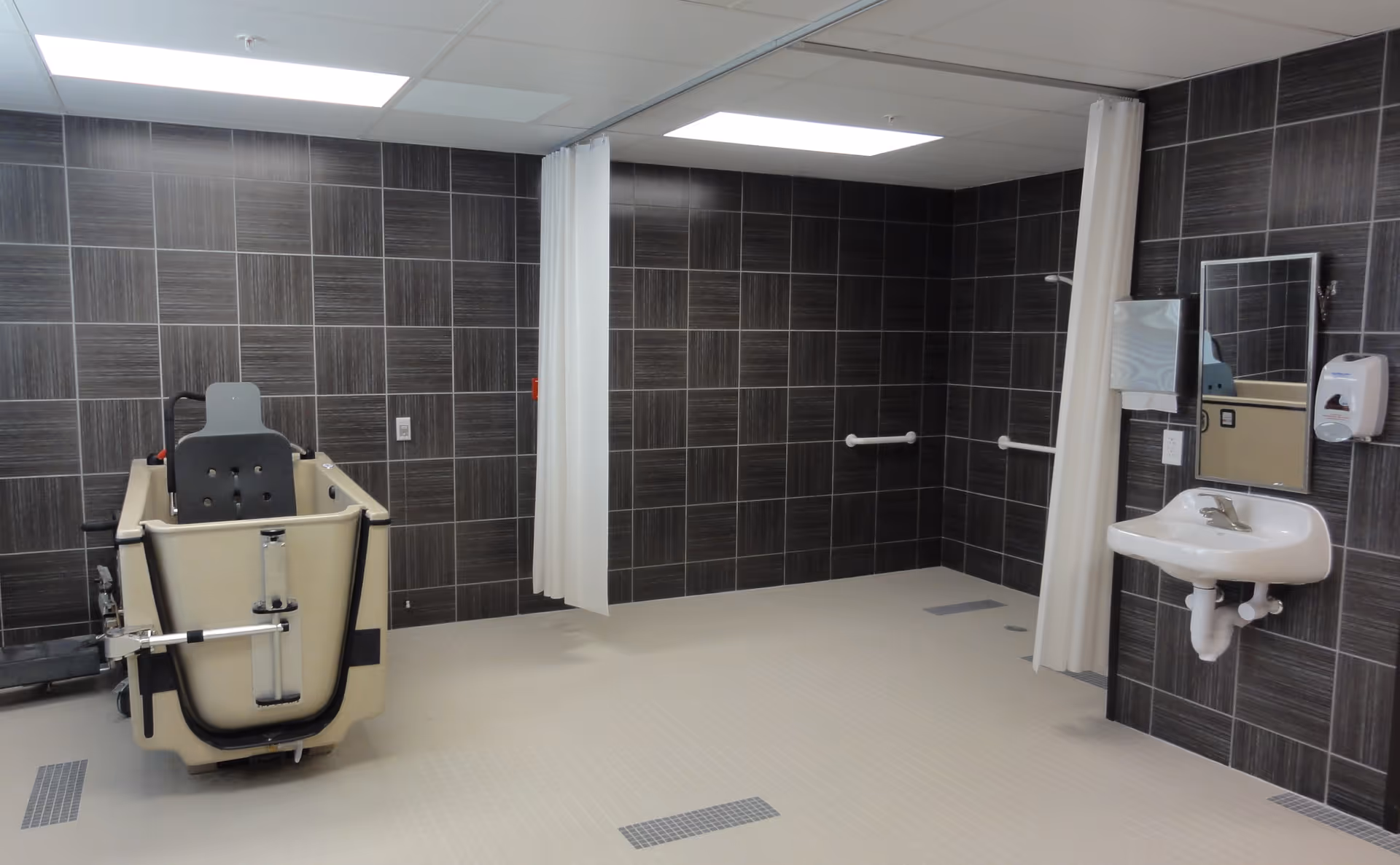 Accessible bathing room with a mobile patient bathtub lift, tiled walls, grab bars, sink, mirror and privacy curtains.