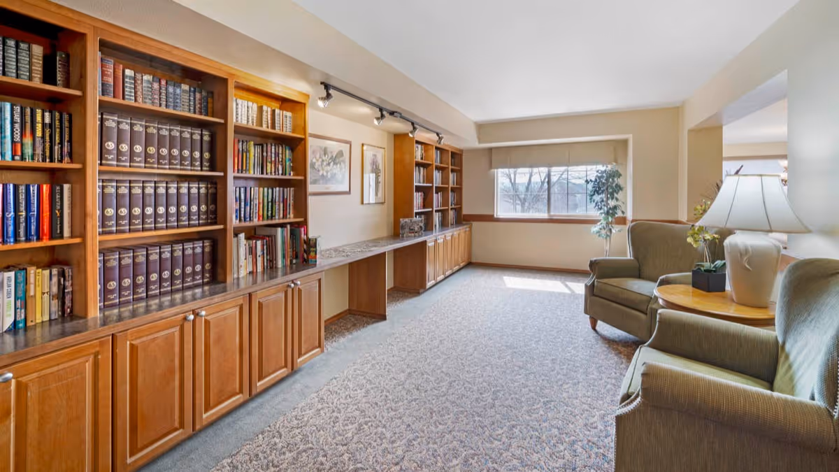 A bright and spacious room with built-in wooden bookshelves filled with books along one wall, a long countertop beneath the shelves, two framed floral artworks on the wall, and two green upholstered armchairs with a round wooden side table and a lamp near a large window letting in natural light.
