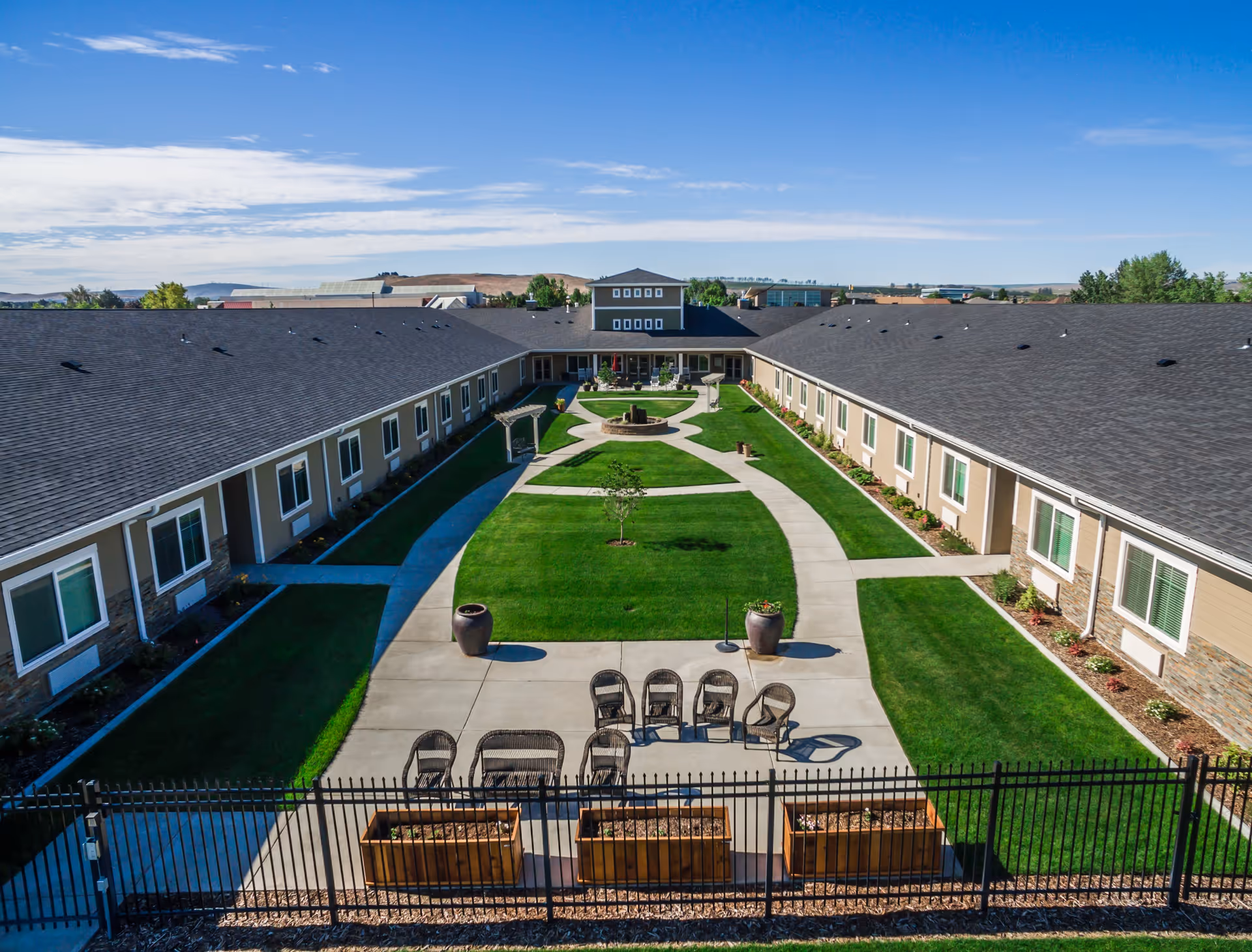 Aerial view of a senior living facility courtyard with a large green lawn, paved walkways, outdoor seating, and a central fountain. The courtyard is surrounded by a single-story building with multiple windows and a dark roof under a clear blue sky.