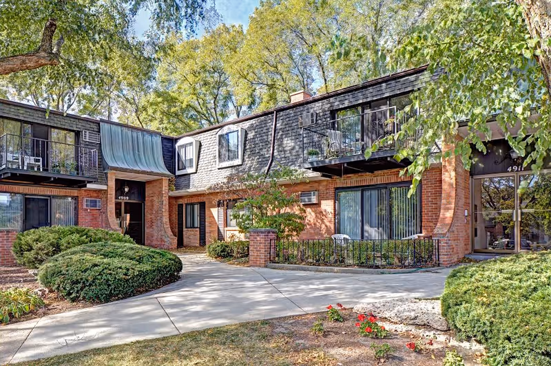 Exterior view of Whitcomb Square Apartments showing a two-story brick building with balconies and large windows surrounded by trees and shrubs on a sunny day.