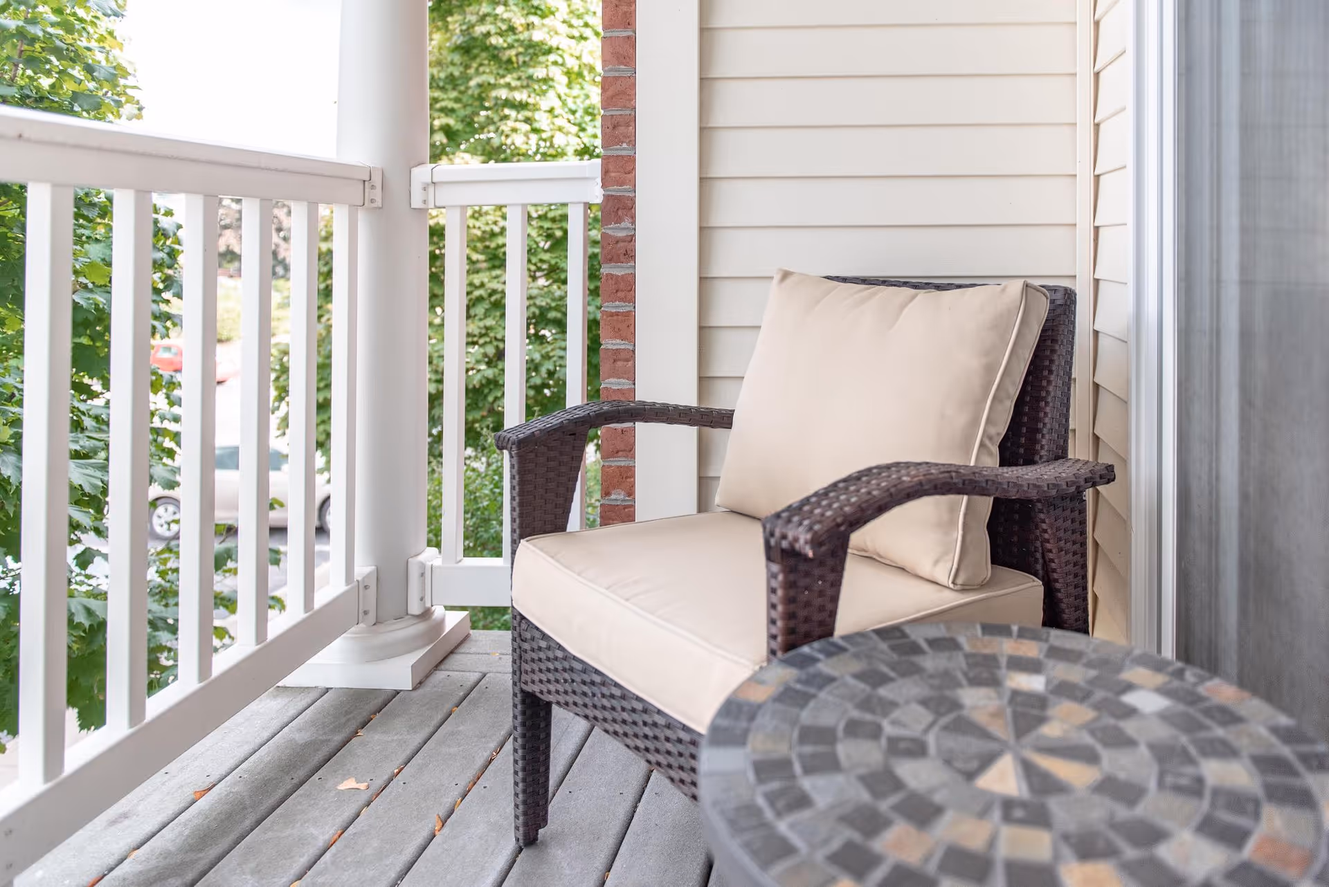 A cushioned wicker chair with beige cushions sits on a wooden balcony next to a round mosaic table. The balcony has white railings and is surrounded by green trees.