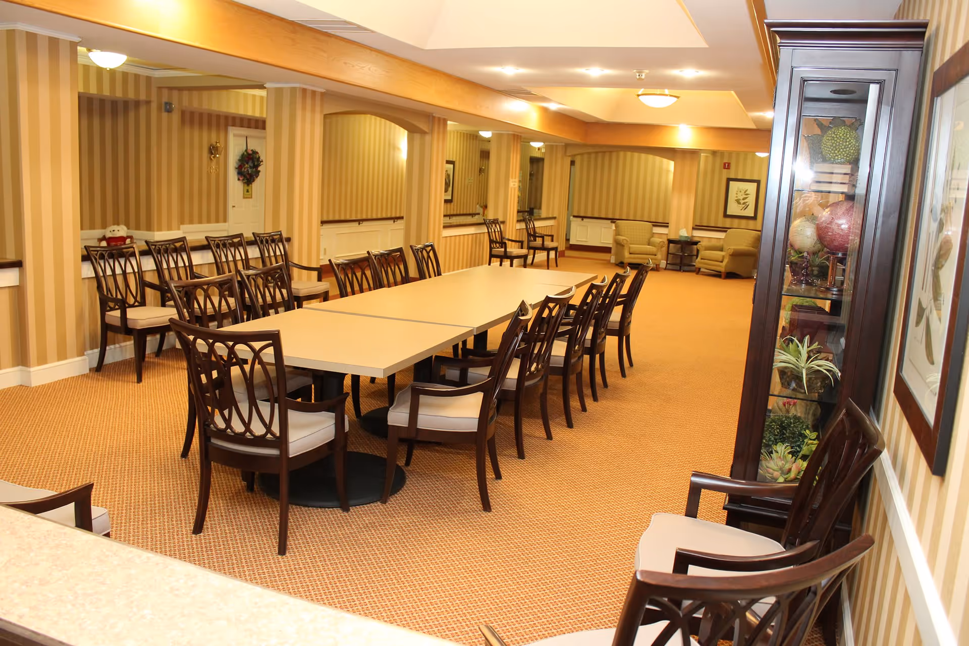 A spacious dining room with a long rectangular table surrounded by wooden chairs with cushioned seats. The room has beige striped wallpaper, carpeted floor, and wooden beams on the ceiling. There are additional chairs along the walls and a glass display cabinet with decorative items on the right side. In the background, there are two armchairs with a small table between them and framed artwork on the walls.