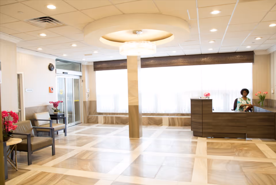 Bright and spacious reception area of a healthcare facility with a woman sitting behind a wooden reception desk. The room features large windows with sheer curtains, a chandelier on the ceiling, beige tiled flooring, and seating area with chairs and pink flowers.