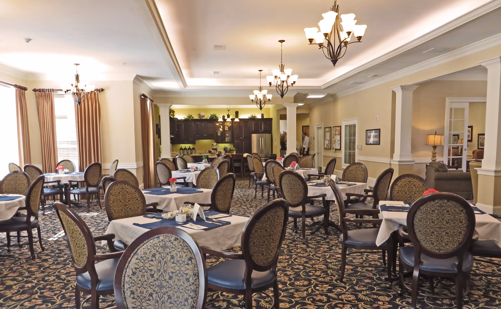 A spacious dining room with multiple round tables covered with white tablecloths and blue placemats, each set with utensils and small flower centerpieces. The room has patterned carpet, beige walls, and several chandeliers hanging from the ceiling. Large windows with brown curtains allow natural light to enter. In the background, there is a kitchen area with dark wood cabinets and stainless steel appliances.
