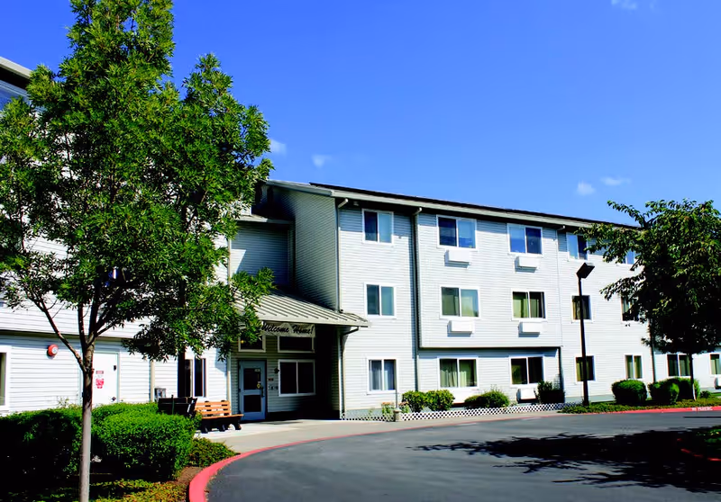 Front exterior of a three-story senior living facility with white siding, a covered entrance, trees, and a curved driveway.