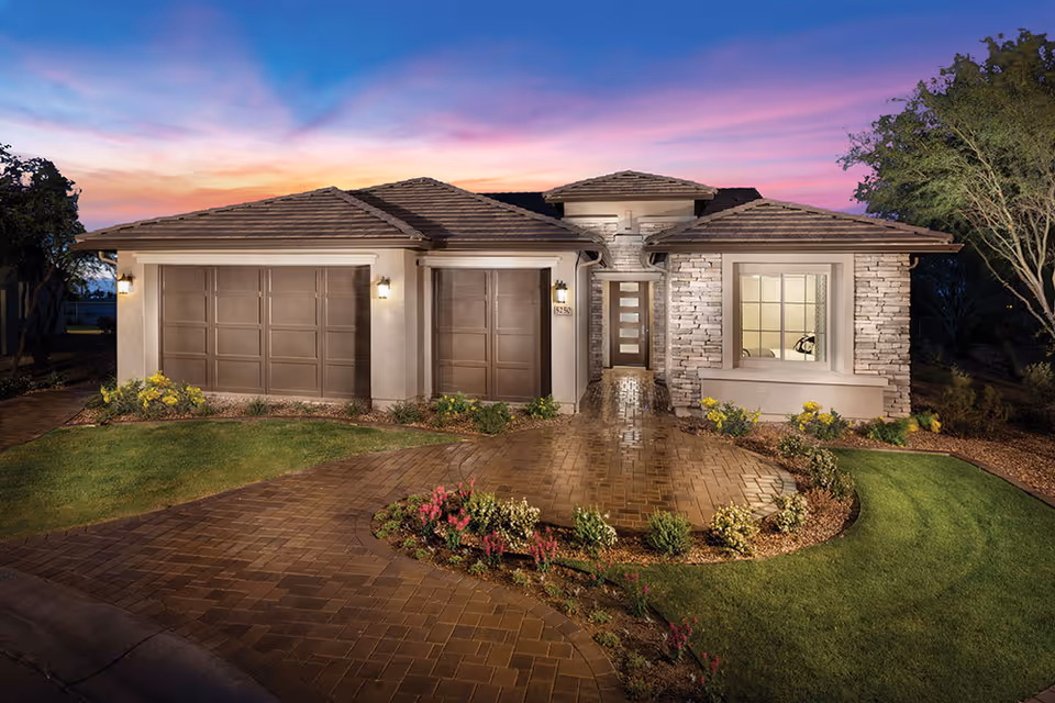 Front exterior of a single-story house with a three-car garage, paved driveway, and landscaped yard at sunset.