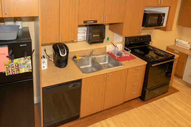 A kitchen area with wooden cabinets, a double stainless steel sink, a black dishwasher, a black electric stove with four burners, a microwave oven mounted above the stove, a black refrigerator with magnets and notes on the door, a coffee maker on the counter, and a dish drying rack with some dishes on a red mat next to the sink.