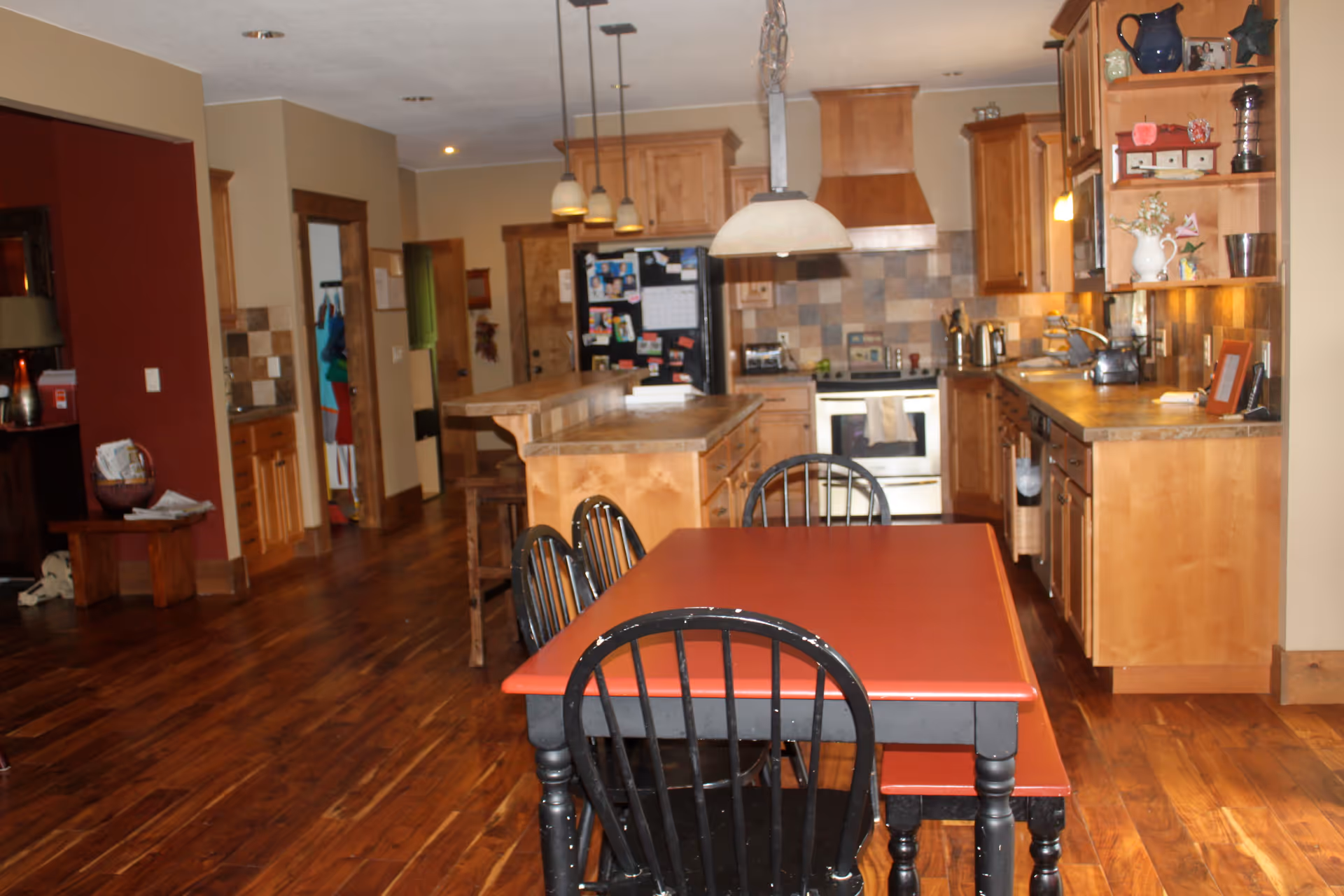 Interior view of a kitchen and dining area with wooden floors and cabinetry. A red dining table with black chairs is in the foreground. The kitchen features a stove, refrigerator with magnets, a kitchen island with stools, and pendant lighting. Various kitchen items and decorations are on the counters and shelves.