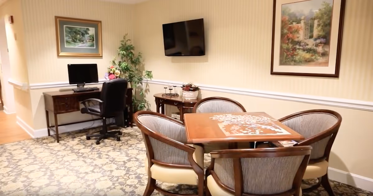 Small common room with a puzzle-covered table surrounded by chairs, a computer desk, wall-mounted TV, and framed artwork.