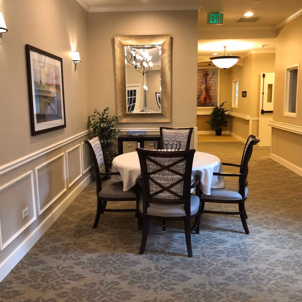 A small seating area in a hallway with a round table covered with a white tablecloth and four cushioned chairs. The walls are beige with white wainscoting, decorated with a framed picture and a large mirror. There are two potted plants, one near the table and one further down the hallway. Ceiling lights illuminate the space, and an exit sign is visible above a doorway in the background.