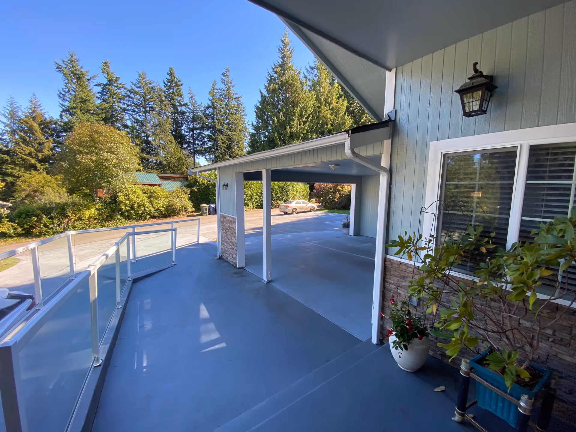 Covered front porch and carport with a ramp, potted plants, and a driveway framed by trees.