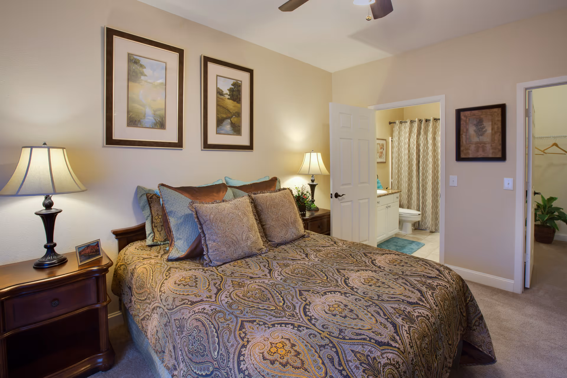 A cozy bedroom with a large bed covered in a patterned bedspread and multiple pillows. Two framed landscape paintings hang above the bed. There are two bedside tables, each with a lamp. An open door reveals a bathroom with a shower curtain, toilet, and sink, while another open door leads to a walk-in closet with a plant inside.
